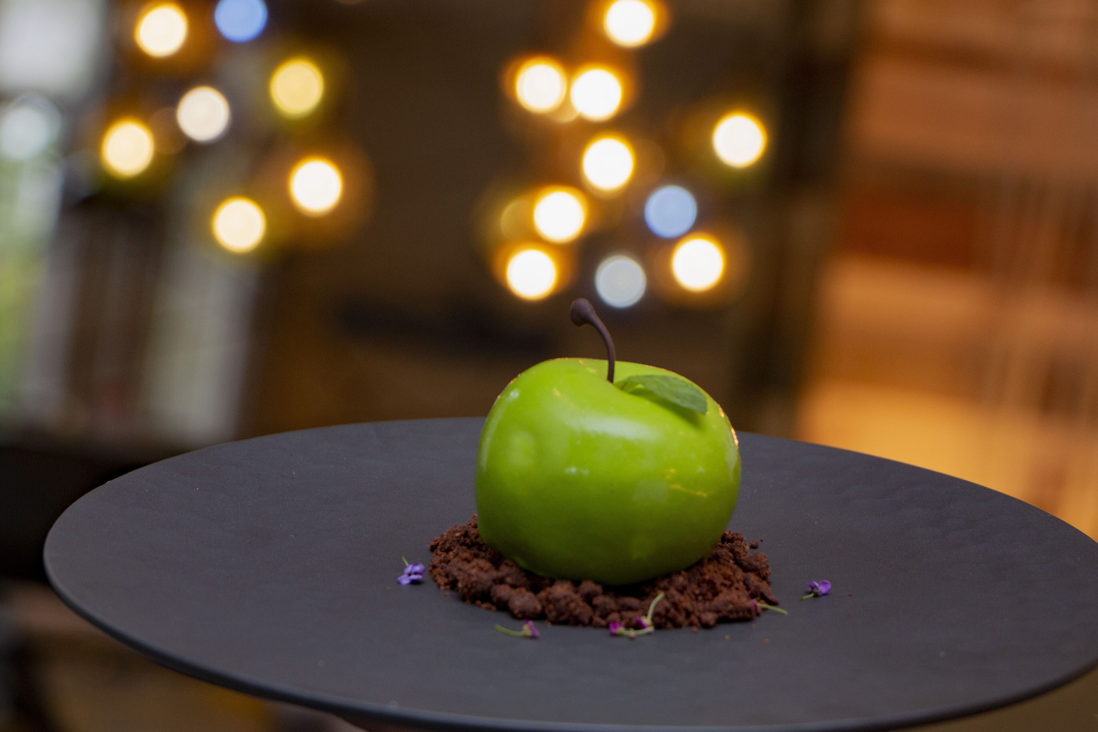 Un postre de manzana verde sobre un plato oscuro, con un fondo borroso de luces.