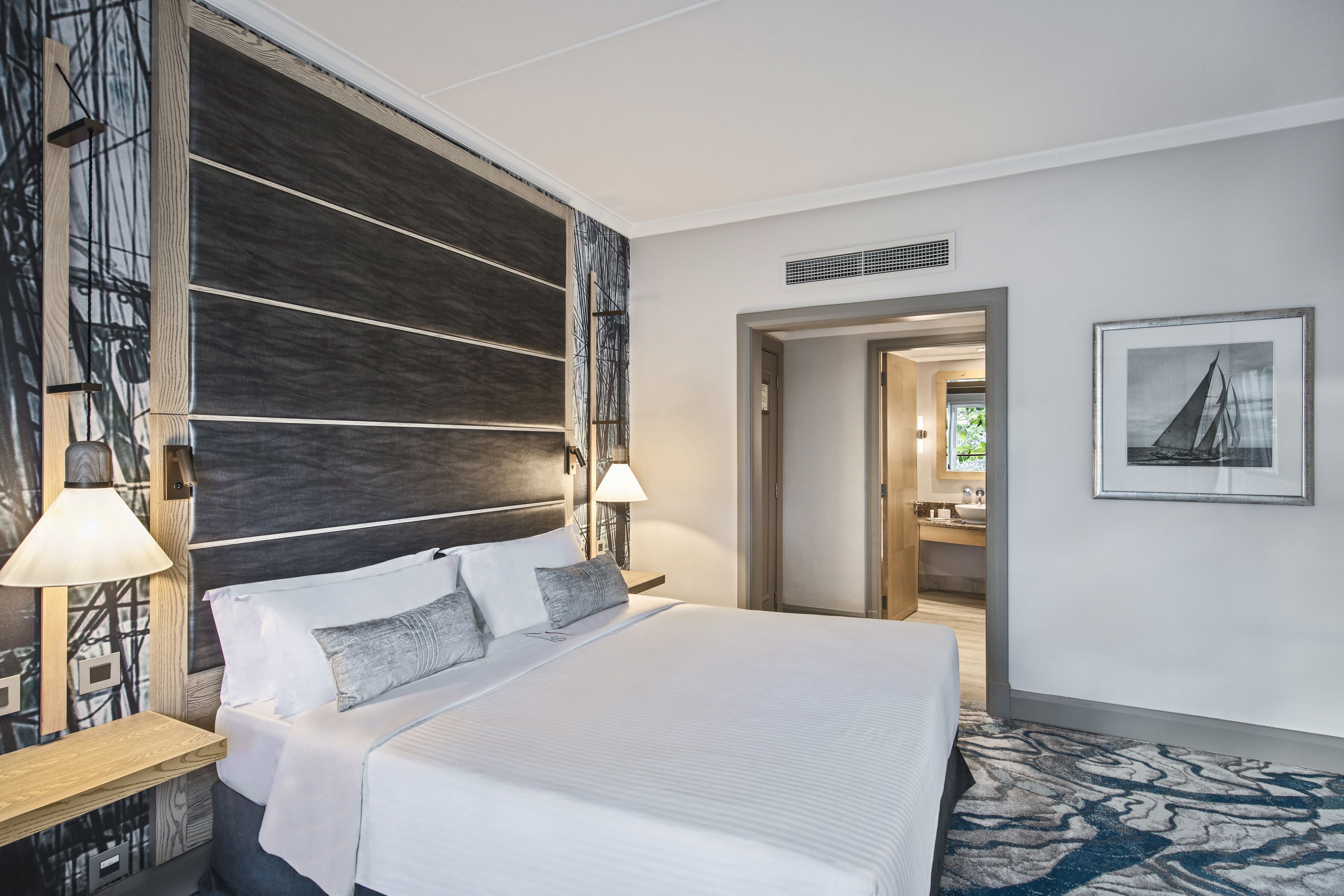 A modern hotel room with a large bed, two bedside lamps, a wooden headboard, a patterned rug, and a framed picture on the wall.