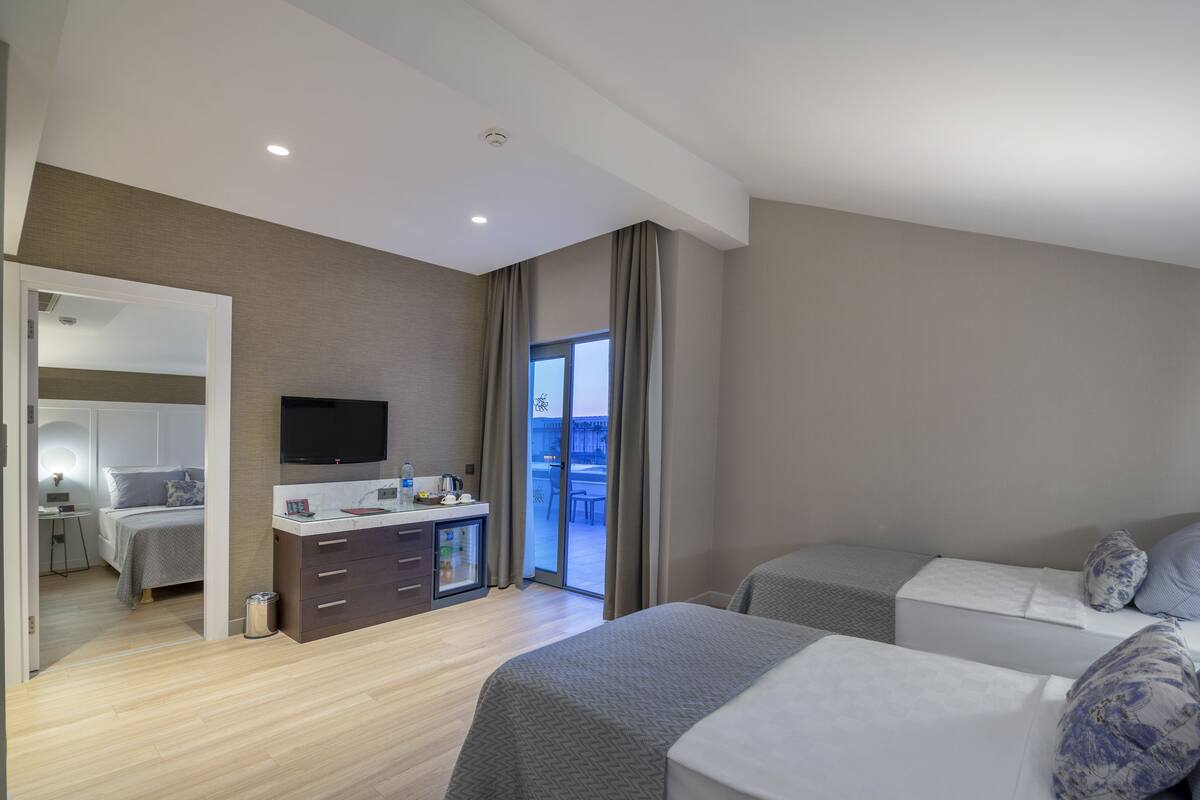 A modern hotel room with two beds, a TV, a small kitchenette, and a balcony with a view.