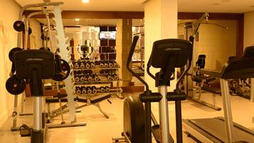 Un gimnasio con máquinas de cardio, equipo de levantamiento de pesas y una pantalla de televisión al fondo.