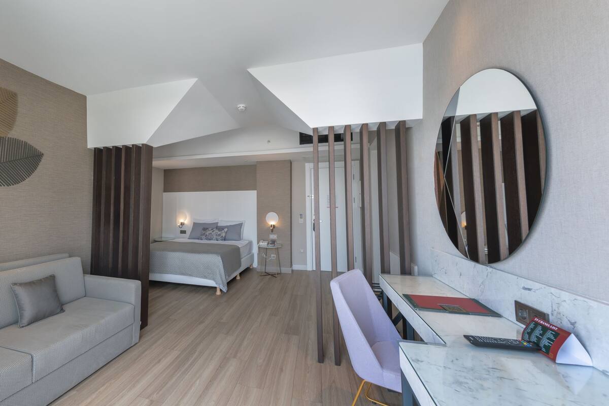 A modern hotel room with a sofa, a bed, a desk with a chair, a large mirror, and a wall-mounted light fixture.