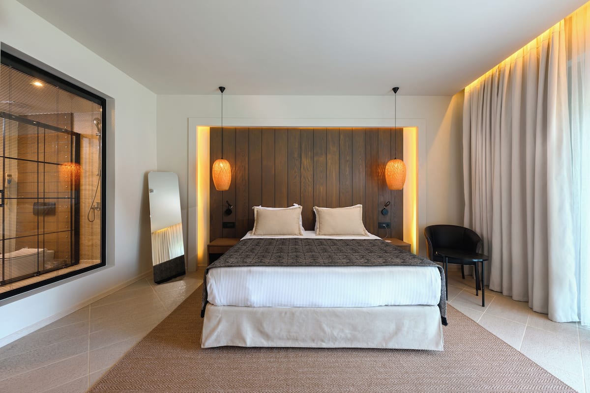 Een moderne hotelkamer met een groot bed, twee nachtkastjes, een spiegel, een stoel en een groot raam met jaloezieën.