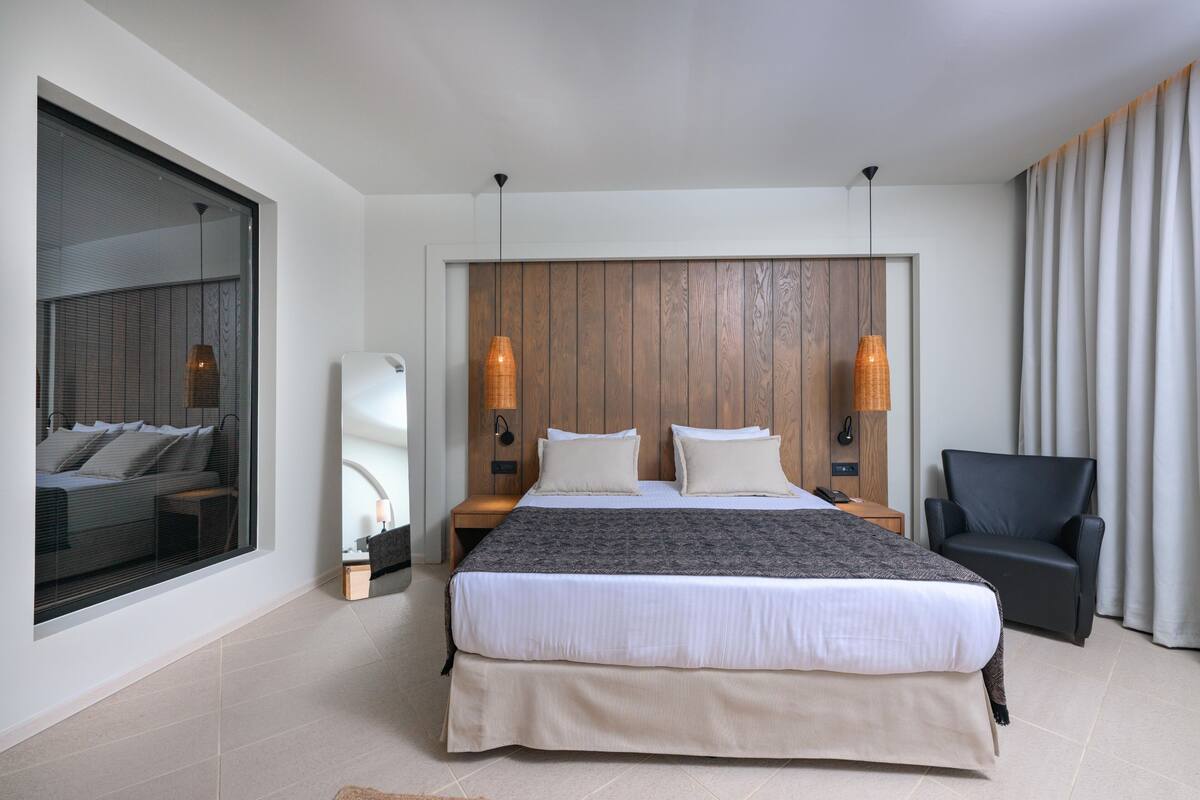 Een moderne hotelkamer met een groot bed, een zithoek met een fauteuil, een groot raam en een houten hoofdbord.