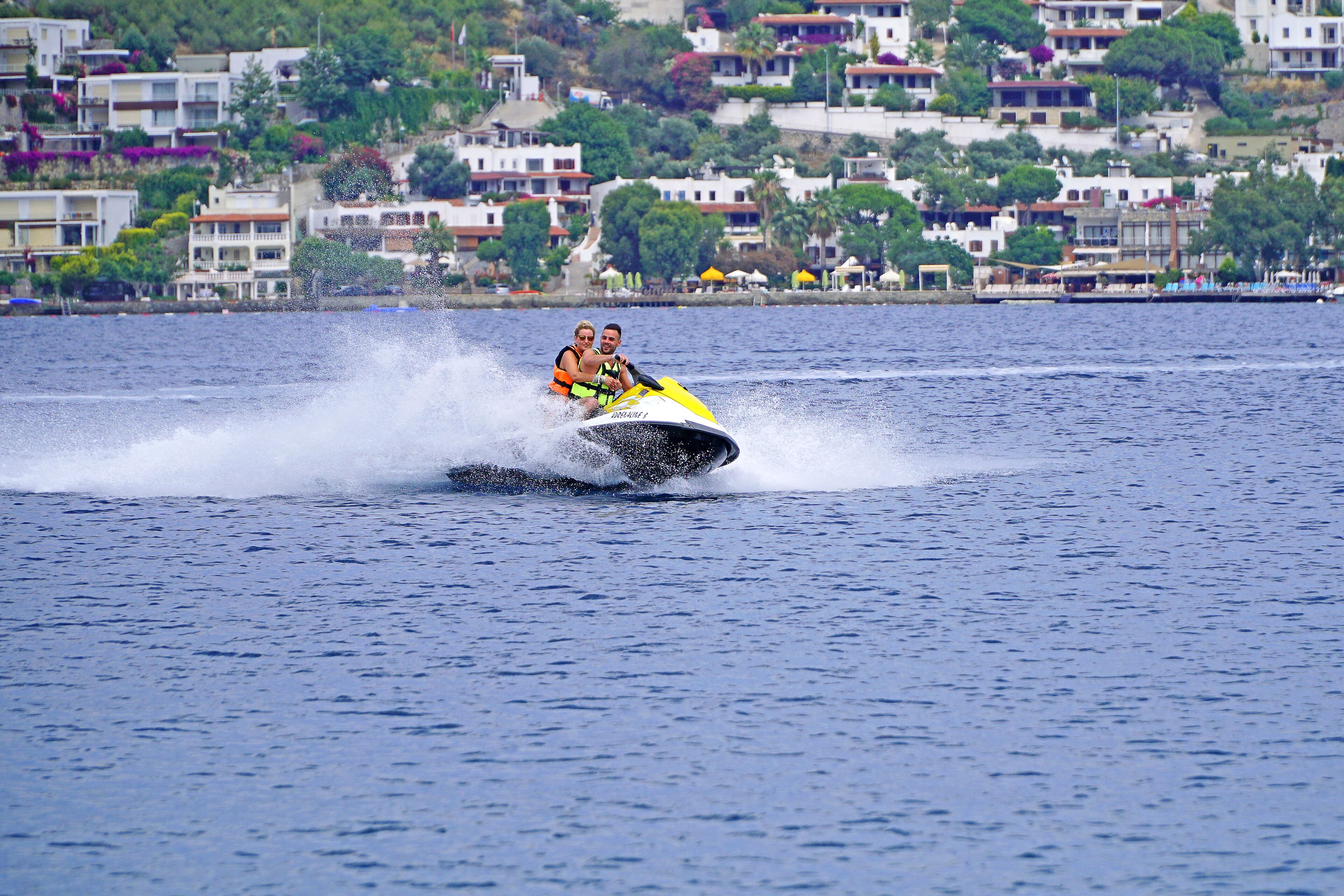 A water jet ski with two passengers in a coastal area with buildings in the background.