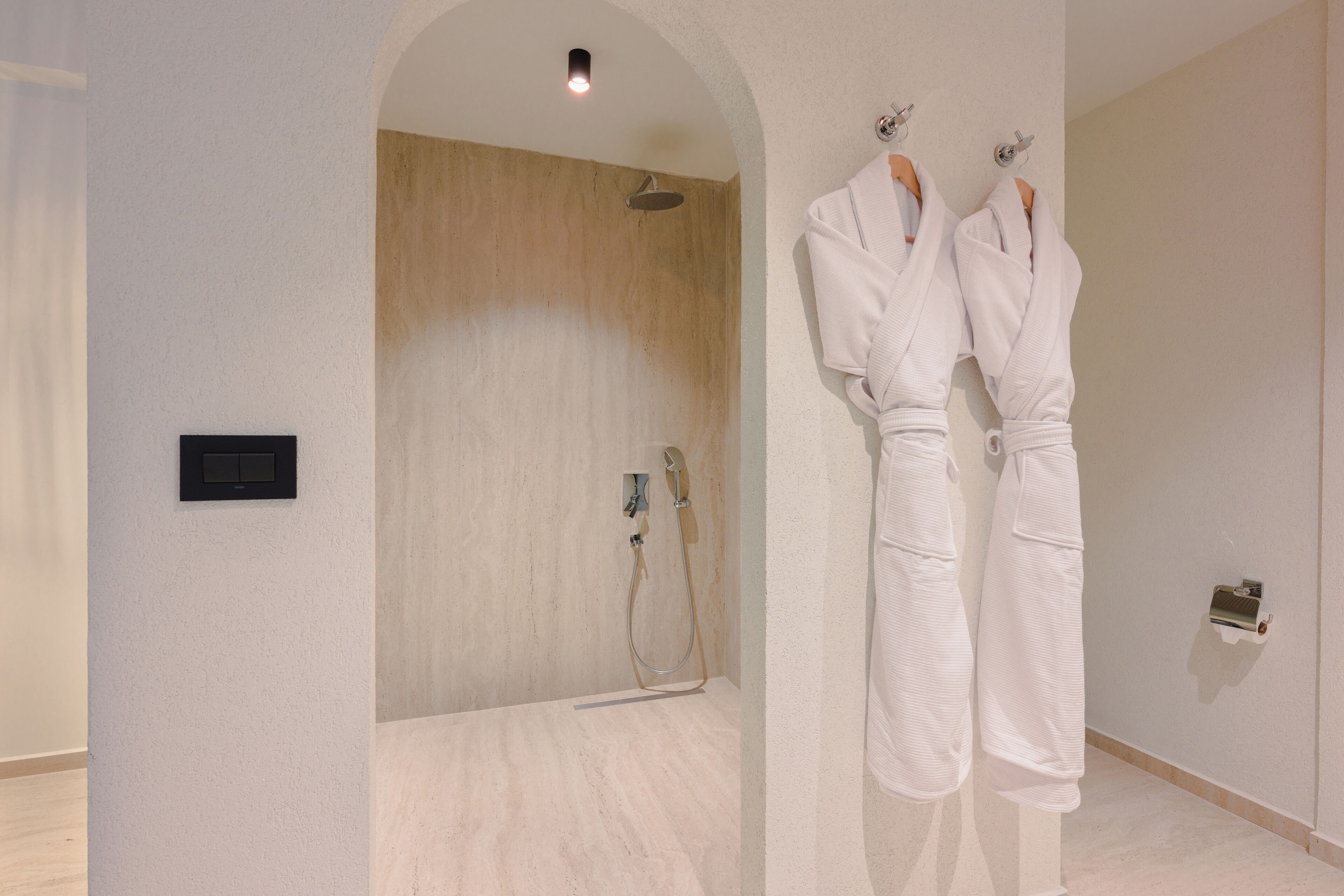 A bathroom with a shower, two white robes hanging on hooks, and a light switch on the wall.