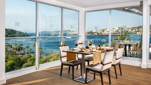 Un coin repas avec une table dressée pour quatre personnes, de grandes fenêtres offrant une vue sur une ville côtière et un port de plaisance.