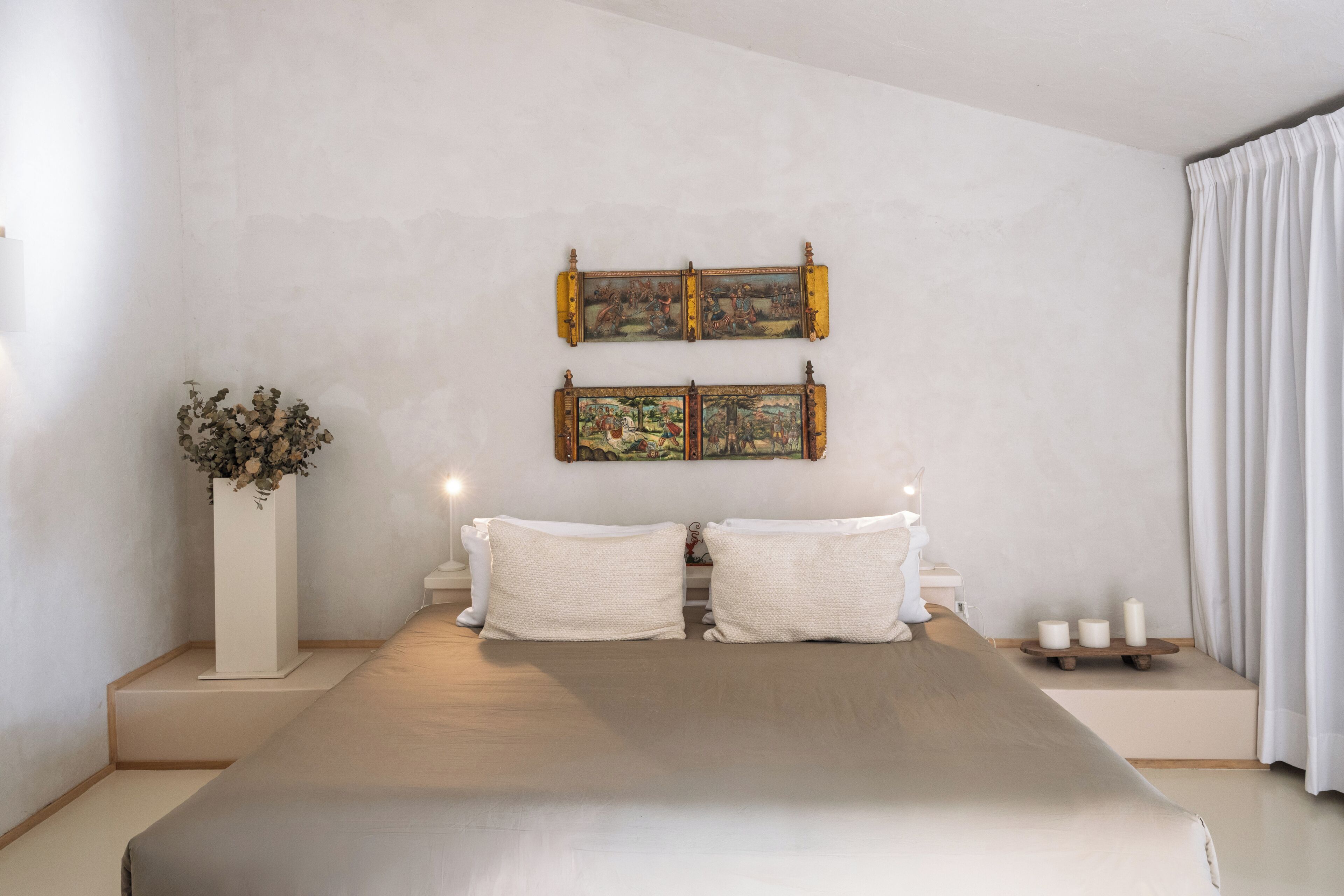 A bedroom with a large bed, two wall-mounted paintings, a white vase with flowers, and a bedside table with candles.