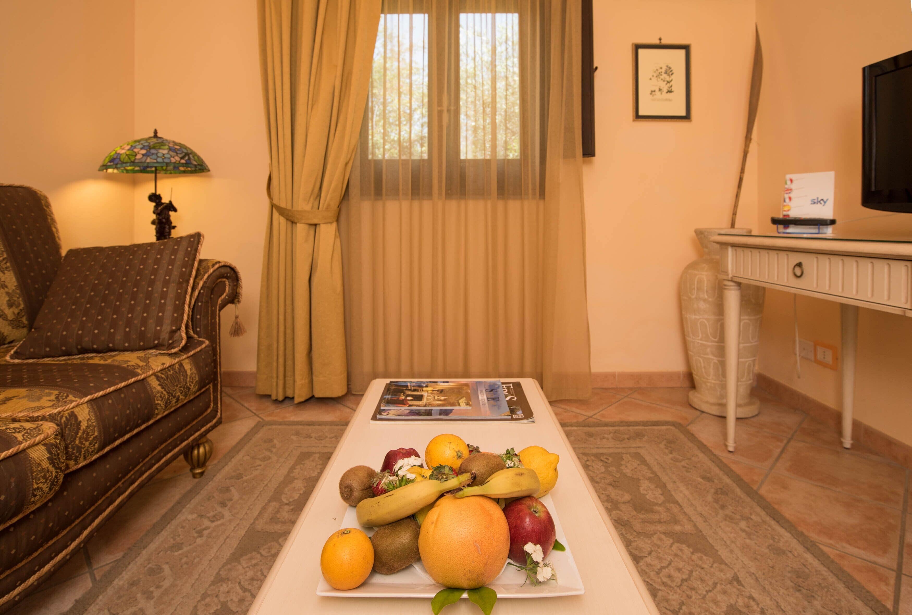 Una sala de estar con un sofá, una mesa de centro con frutas, una lámpara, un televisor y un cuadro en la pared.