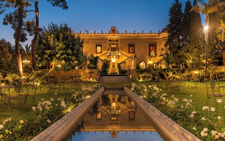 A grand building with a central staircase, surrounded by lush gardens and a reflective water feature.