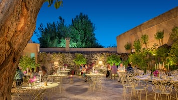 Un espace de restauration en plein air avec des tables et des chaises rondes, entouré de verdure et d’un mur de pierre.