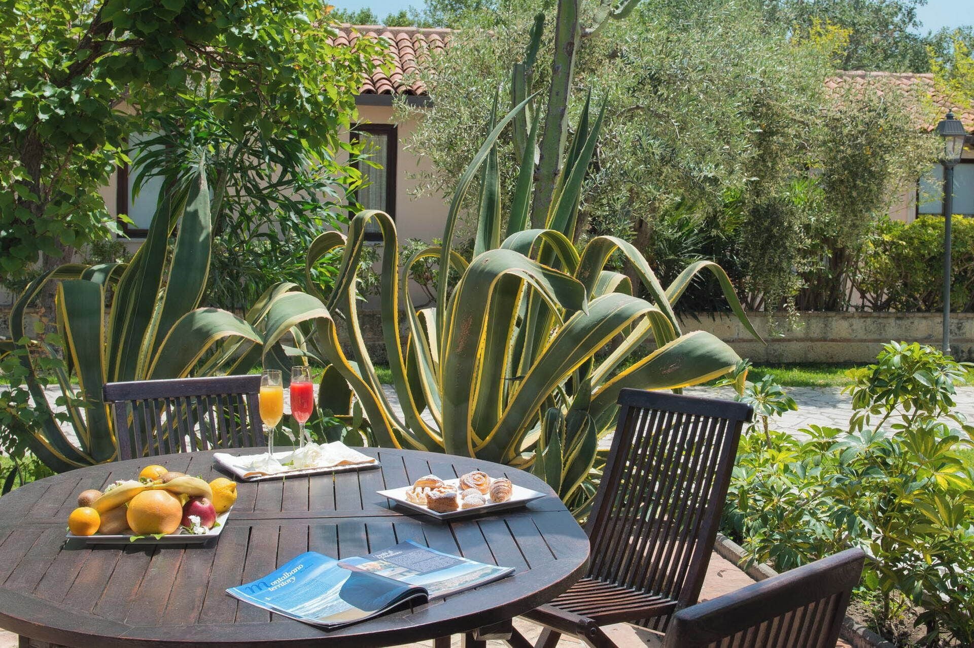 Une table ronde en bois, sur laquelle sont posés un magazine, des fruits et des pâtisseries, entourée de chaises dans un cadre extérieur agrémenté de grandes plantes vertes et d’un bâtiment en arrière-plan.