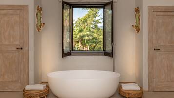 Ein Badezimmer mit einer freistehenden Badewanne, zwei Korbstühlen und einem Fenster mit Blick auf Bäume.