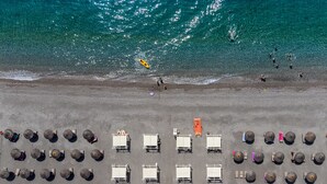 Vista aèria d'una platja amb files de cadiretes i ombrelles, una torre de socorristes i persones a l'aigua.