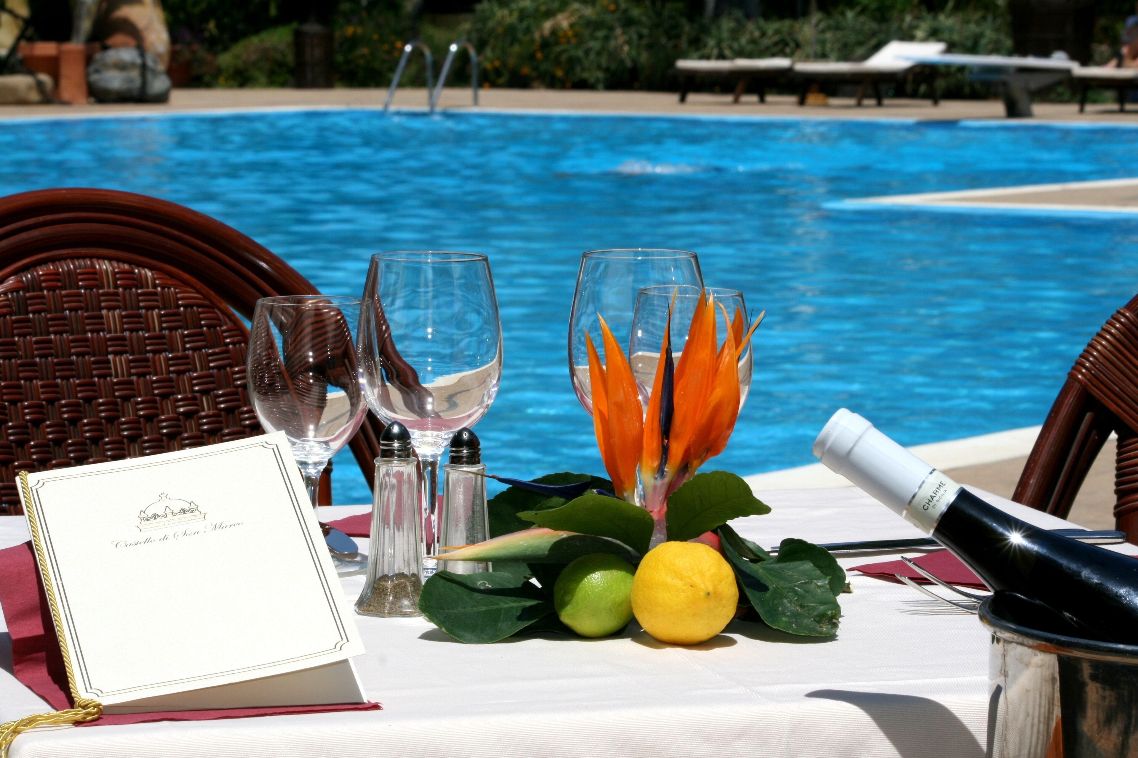 Mesa dispuesta con un menú, copas de vino, una botella de champán y un arreglo decorativo de hojas y cítricos junto a una piscina.