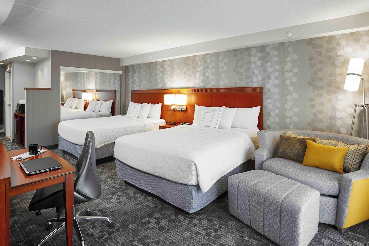 A hotel room with two beds, a desk with a laptop, a chair, a sofa, and a lamp.