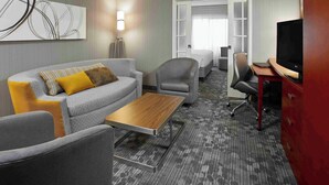 A hotel room with a grey sofa, a wooden coffee table, a TV, and a desk with drawers. - Courtyard by Marriott Toronto Brampton (Brampton)