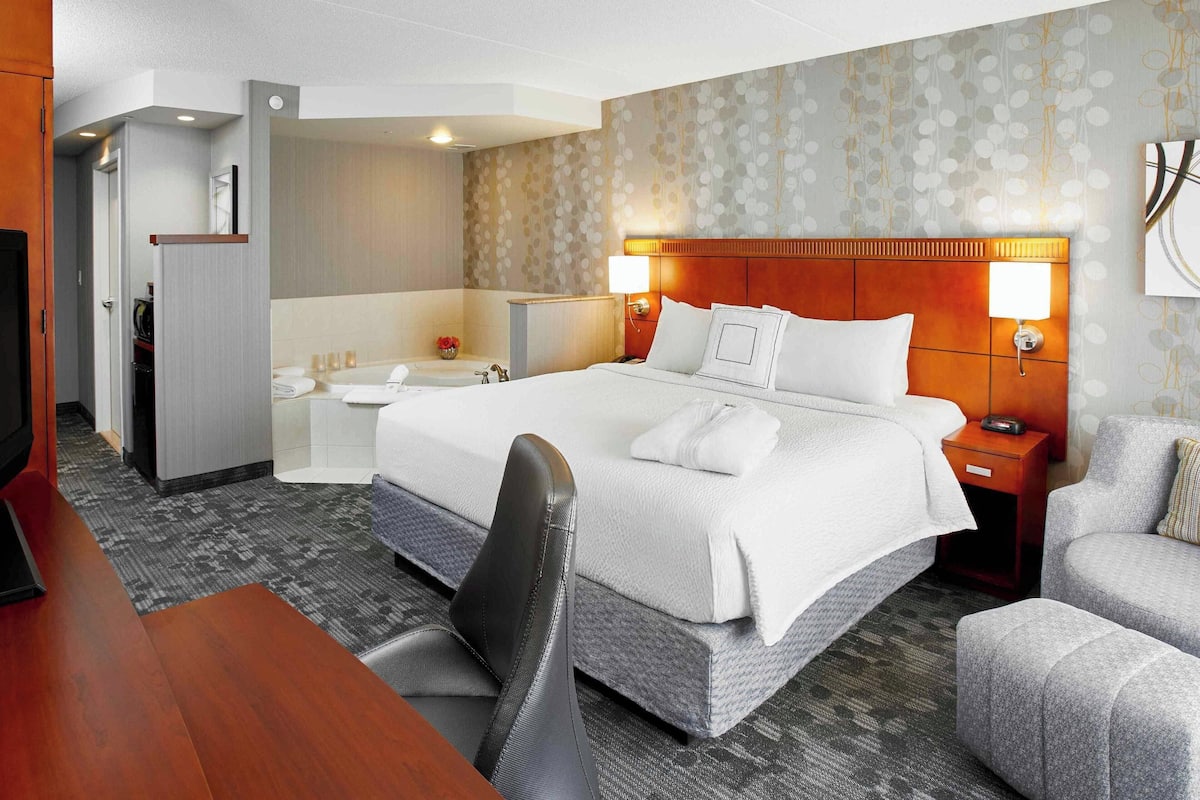 A hotel room with a large bed, a desk, a chair, a small table, a nightstand, and a lamp.