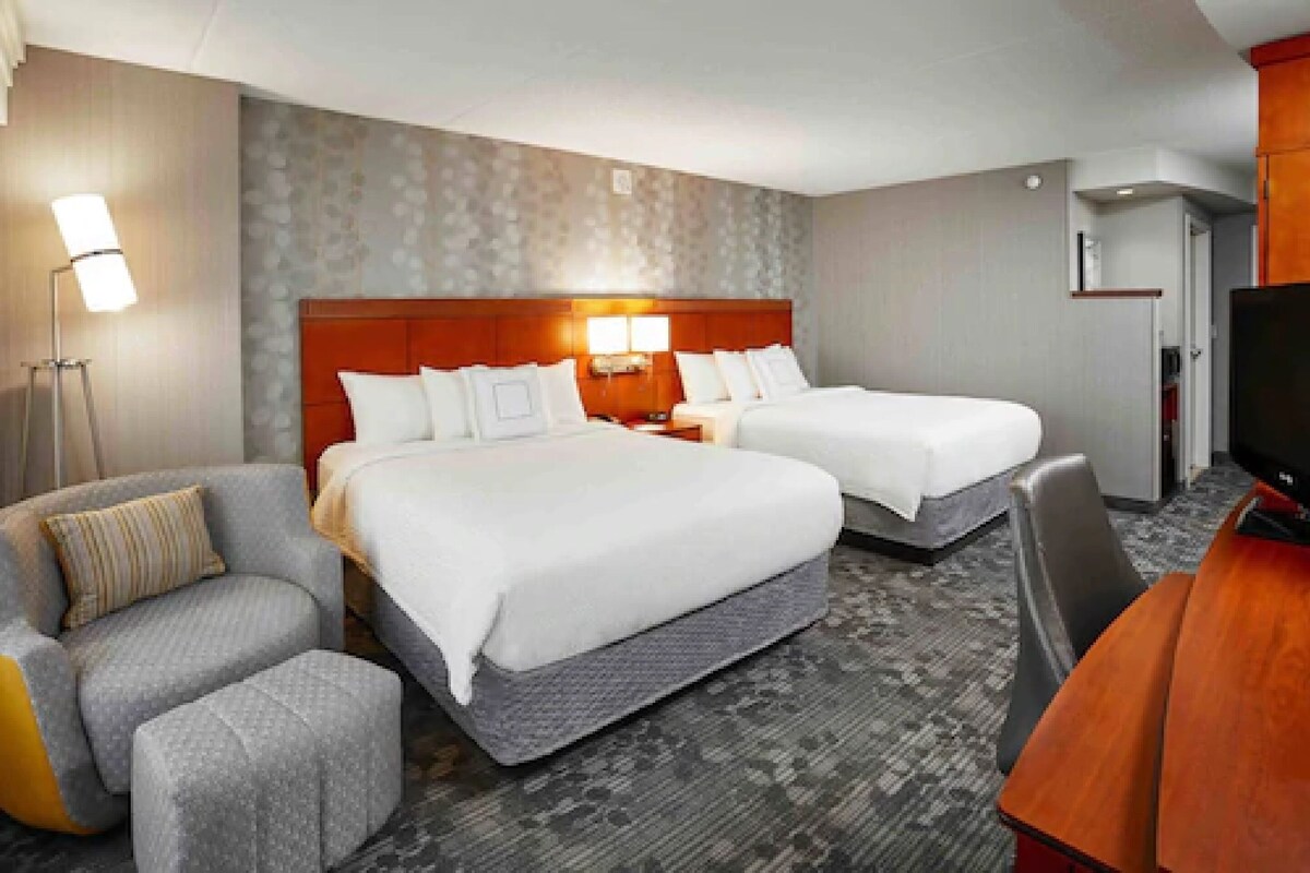 A hotel room with two beds, a desk, a chair, and a TV.