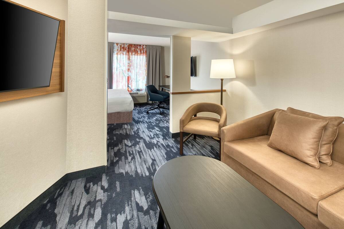 A hotel room with a sofa, a TV mounted on the wall, a desk with a lamp, and a view of the hallway.
