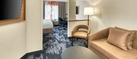 A hotel room with a sofa, a TV mounted on the wall, a desk with a lamp, and a view of the hallway.