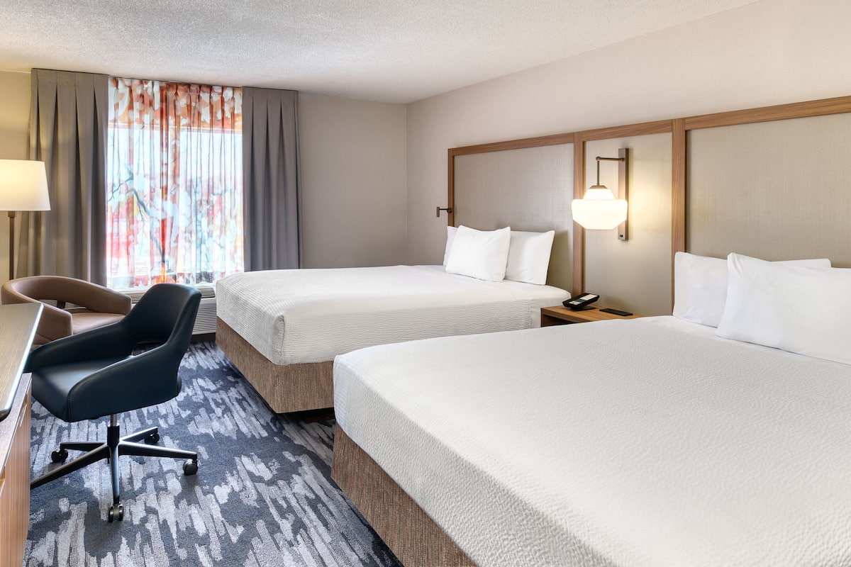 A hotel room with two beds, a desk with a chair, a lamp, and a window with curtains.