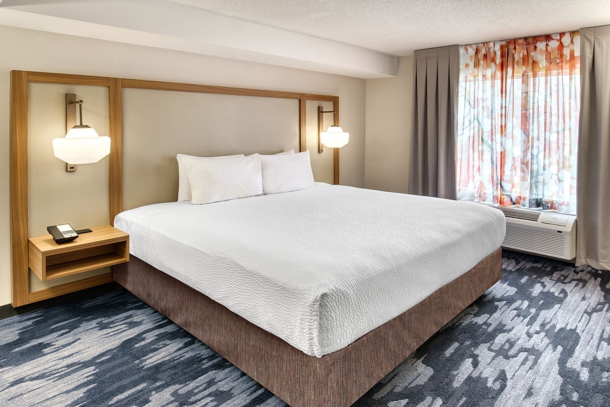 A hotel room with a large bed, wooden headboard, two bedside lamps, a small bedside table with a phone, and a window with curtains.