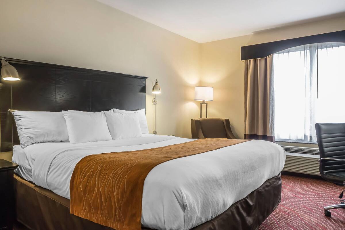 Hotellihuone, jossa on suuri sänky, kaksi yöpöytävalaisinta, tuoli ja ikkuna, jossa on verhot.