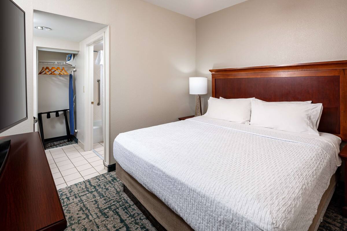 A hotel room with a large bed, a wooden headboard, a bedside table with a lamp, a wardrobe, and a television.