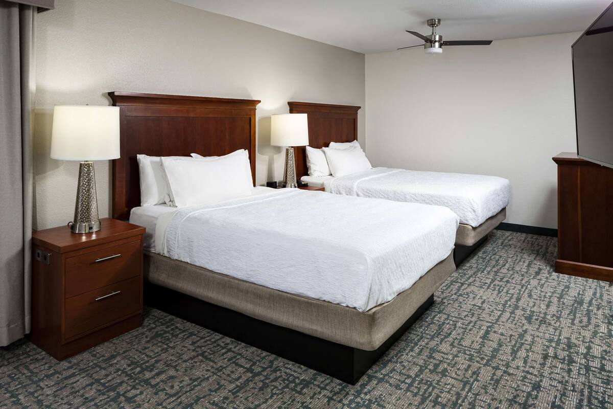 A hotel room with two beds, a wooden headboard, a nightstand with a lamp, and a carpeted floor.