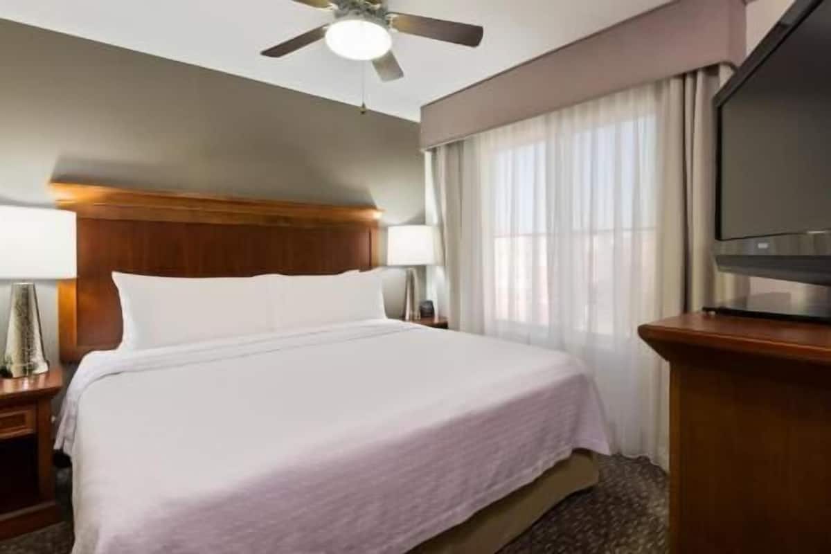 A hotel room with a bed, a ceiling fan, a TV, and a window with curtains.