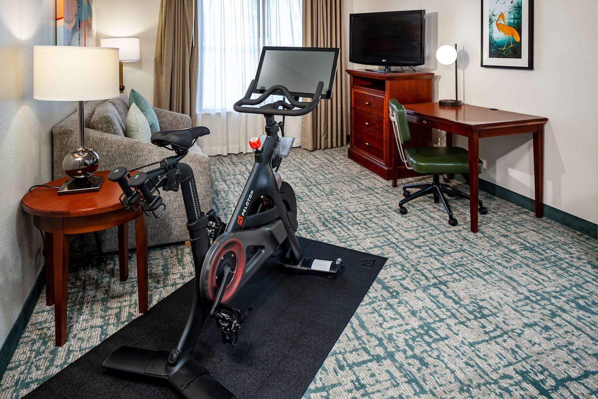 A hotel room with a stationary bike, a desk, a television, a lamp, and a couch.