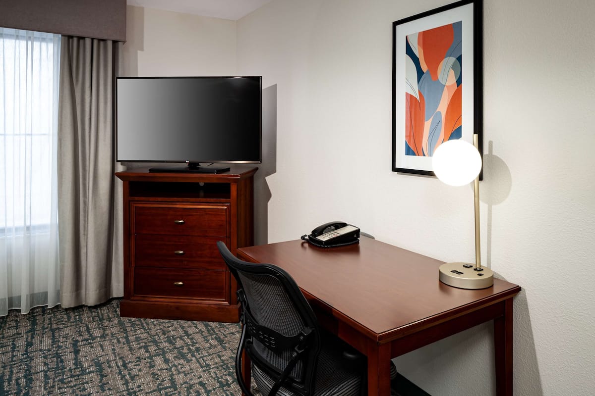 A hotel room with a wooden desk, a black office chair, a television on a wooden stand, a telephone, and a framed abstract painting on the wall.