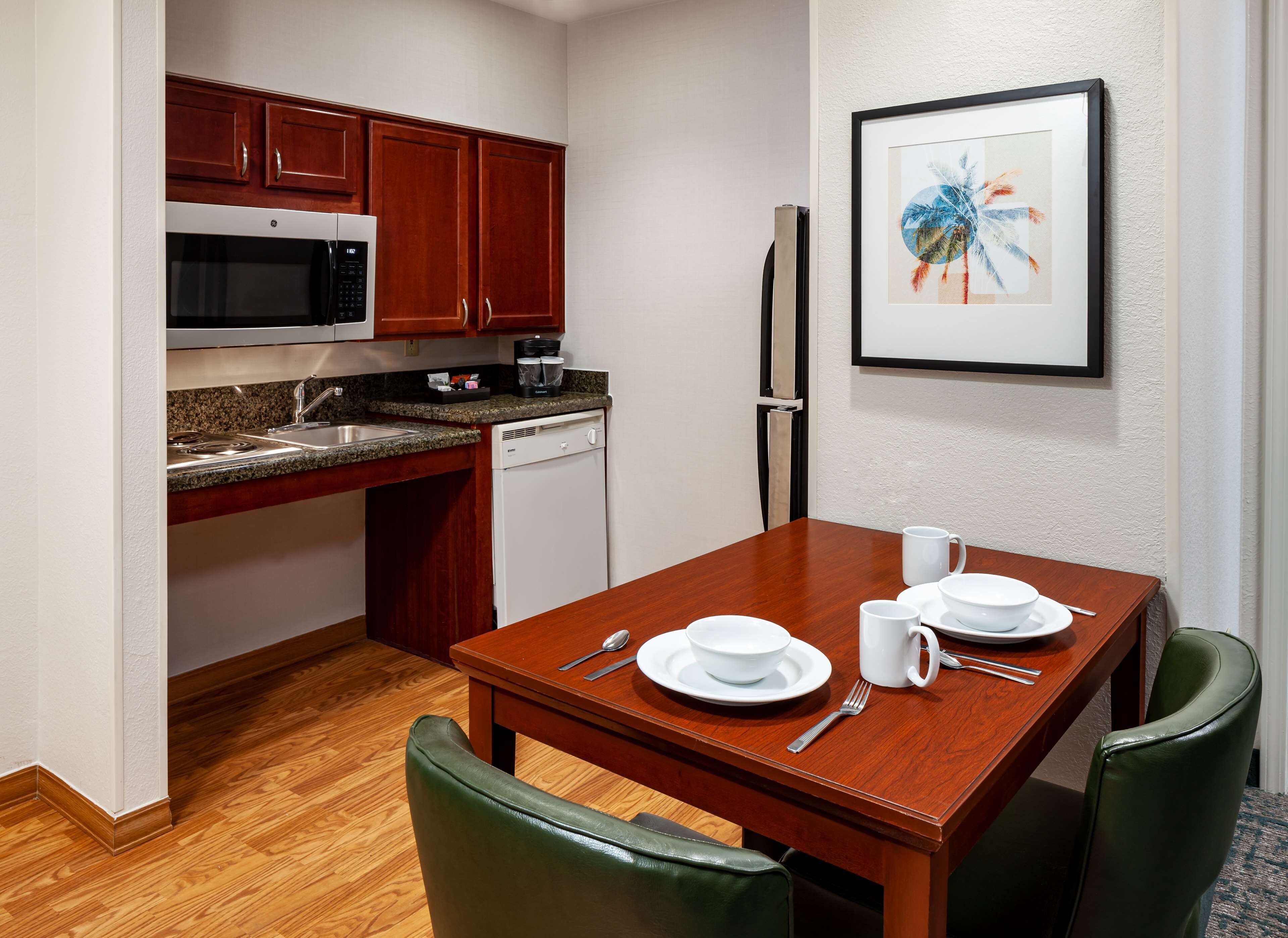 A compact kitchen with a microwave, coffee maker, and dishwasher. A dining table with white plates, cups, and cutlery is set.