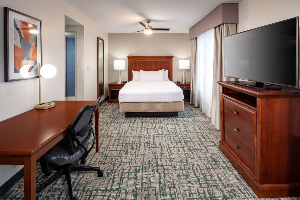 A hotel room with a large bed, a desk with a chair, a television, and a ceiling fan.