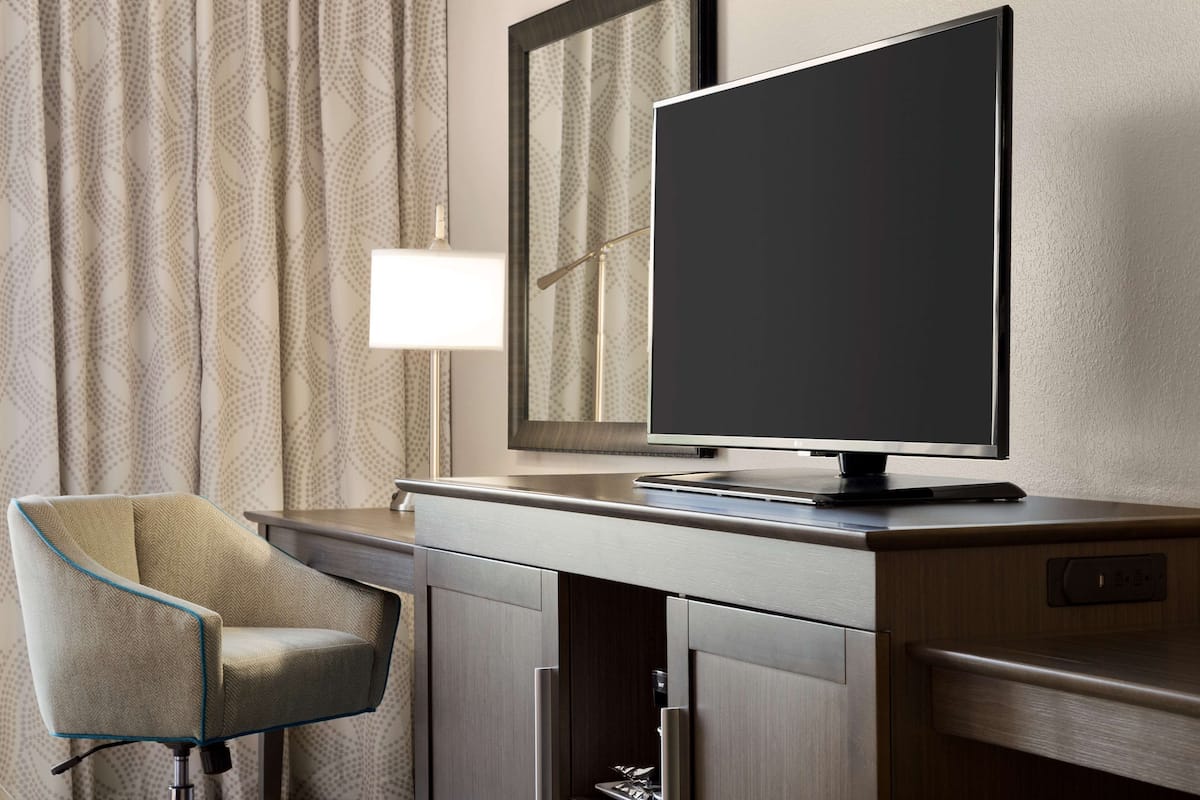 A modern hotel room with a flat-screen TV on a wooden desk, a chair with a blue trim, and a large mirror above the desk.