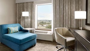 Habitación de hotel con sofá azul, sillón beige, escritorio y ventana grande con vista a la ciudad.