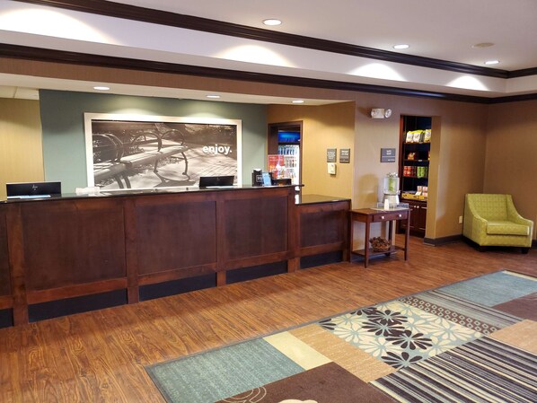 A hotel reception area with a wooden reception desk, a large poster, a small table with a chair, and a minibar.