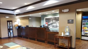 Zona de recepción de hotel con mostrador de madera, minibar, estación de café y refrigerador con bebidas.