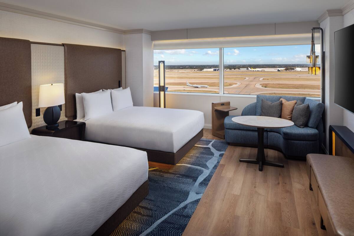A hotel room with two beds, a sofa, a small table, and a view of an airport.