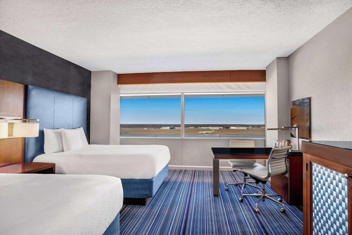 Habitación de hotel con dos camas, un escritorio, una silla y un ventanal con vistas al aeropuerto.
