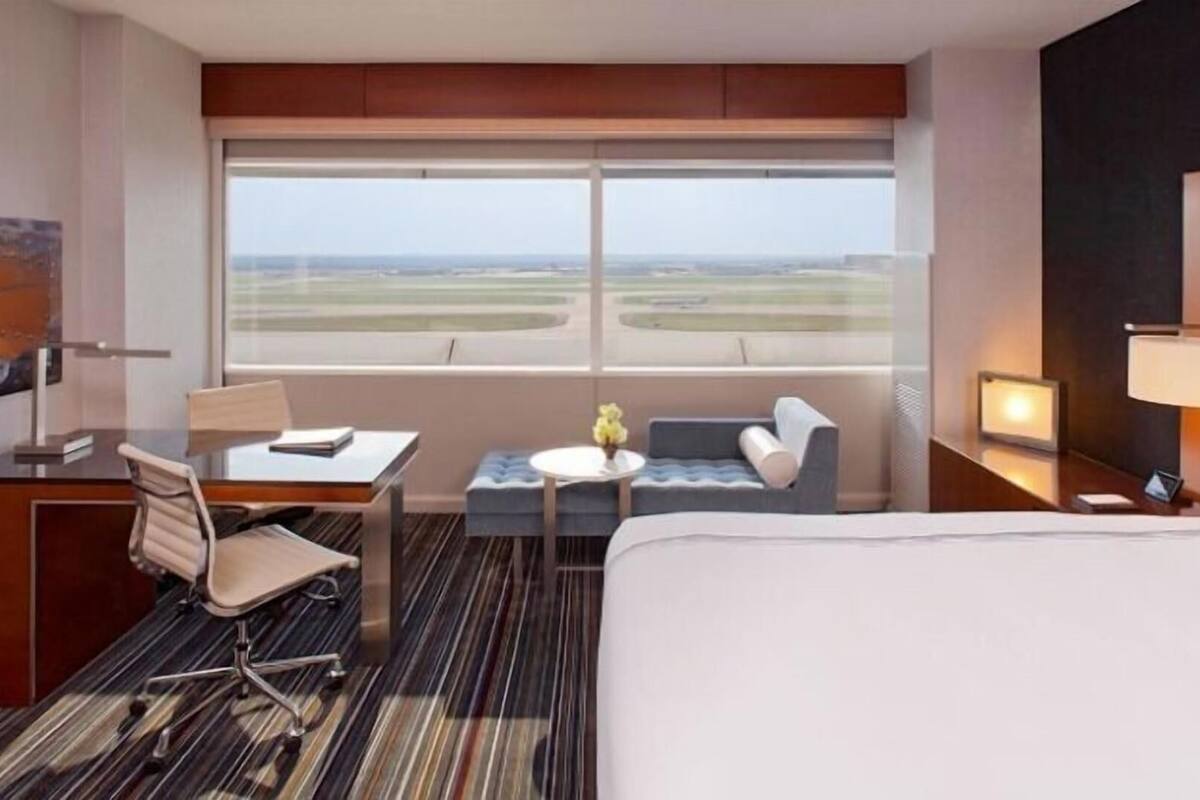 A hotel room with a bed, a desk, a chair, a small table with a vase, and a large window with a view of an airport.