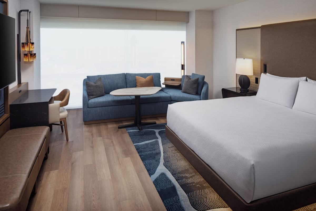 A hotel room with a large bed, a sofa, a small table, a TV, and a nightstand with a lamp.