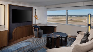 Habitación de hotel moderna con una televisión grande, un escritorio, un sofá cómodo y vistas al aeropuerto.