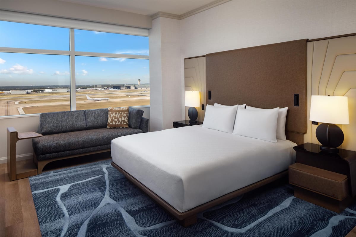 A hotel room with a large bed, a sofa, a nightstand with lamps, and a view of an airport.
