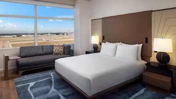 A hotel room with a large bed, a sofa, a nightstand with lamps, and a view of an airport.