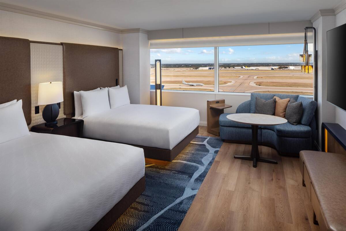 A hotel room with two beds, a sofa, a small table, and a large window with an airport view.