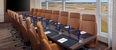 Un salón de conferencias con una gran mesa, sillas marrones, un televisor de pantalla plana y vistas al aeropuerto.