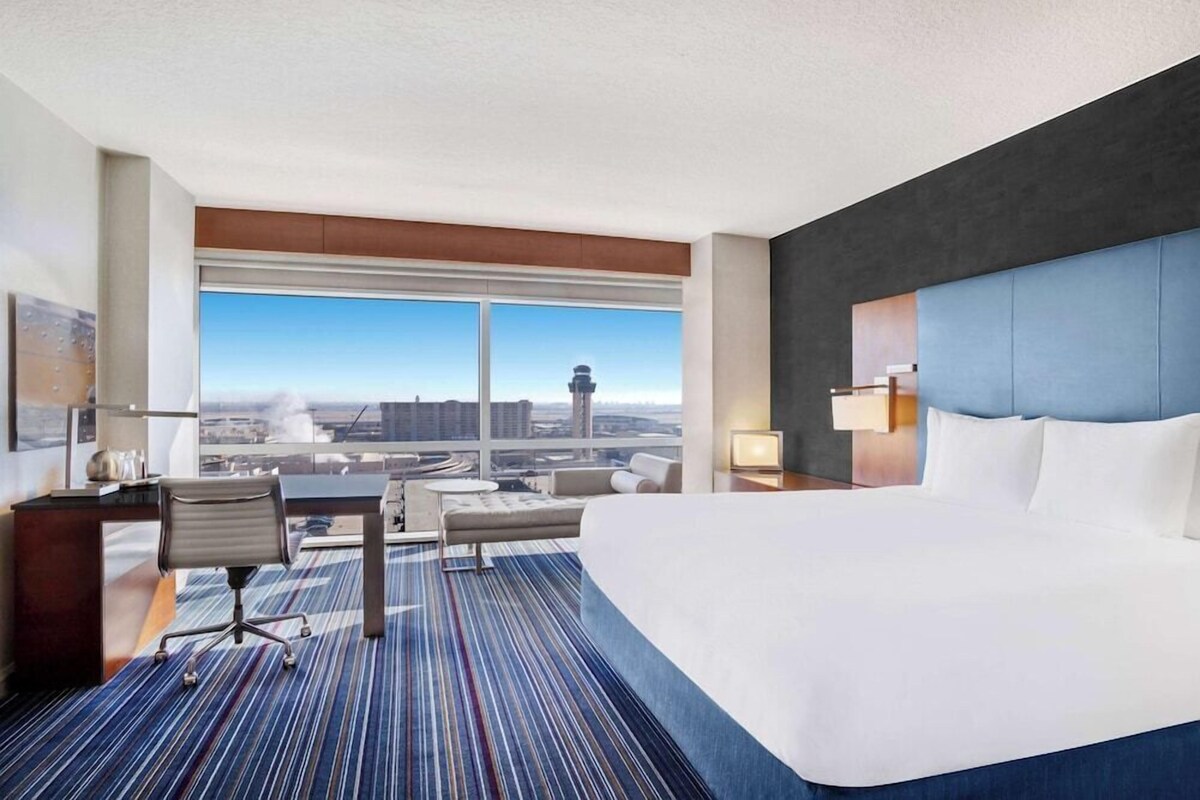Habitación de hotel con una cama grande, un escritorio, una silla y vistas a la ciudad a través de un ventanal amplio.