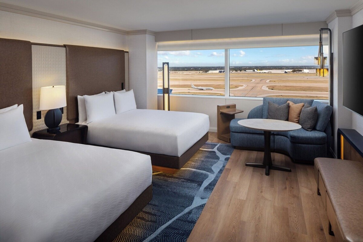 Habitación de hotel con dos camas, un sofá, una mesita y un ventanal con vista al aeropuerto.