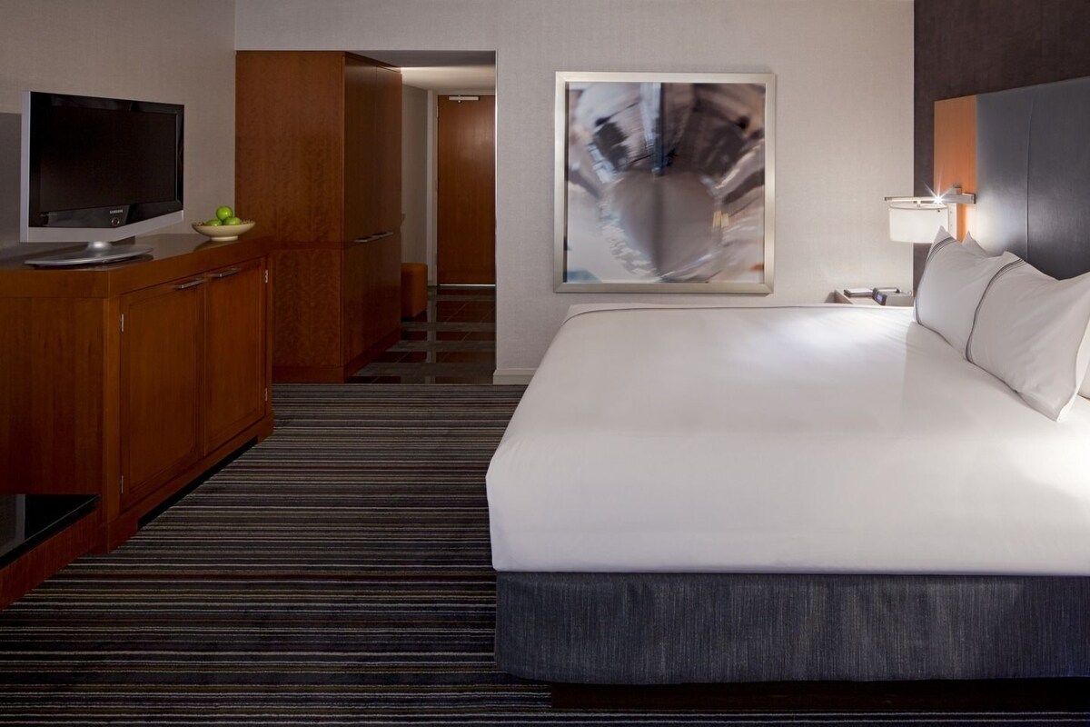 Habitación de hotel con cama, televisor, escritorio y un frutero, además de un cuadro en la pared.
